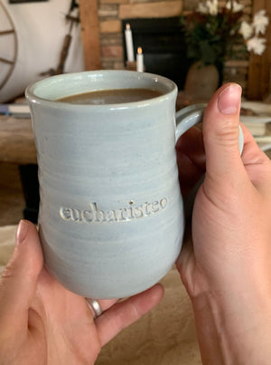 Three Gratitude Mugs with unique designs and glaze colors: "eucharisteo" with cloud blue glaze on white clay, "give thanks always" with darker blue glaze and ochre undertones, and "be still" with variegated gray glaze on red clay. Elegant shape, slight ripples, curved handles. Perfect for reflection and gifting.