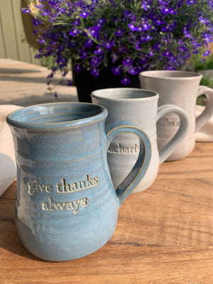 Three Gratitude Mugs with unique designs and glaze colors: "eucharisteo" with cloud blue glaze on white clay, "give thanks always" with darker blue glaze and ochre undertones, and "be still" with variegated gray glaze on red clay. Elegant shape, slight ripples, curved handles. Perfect for reflection and gifting.