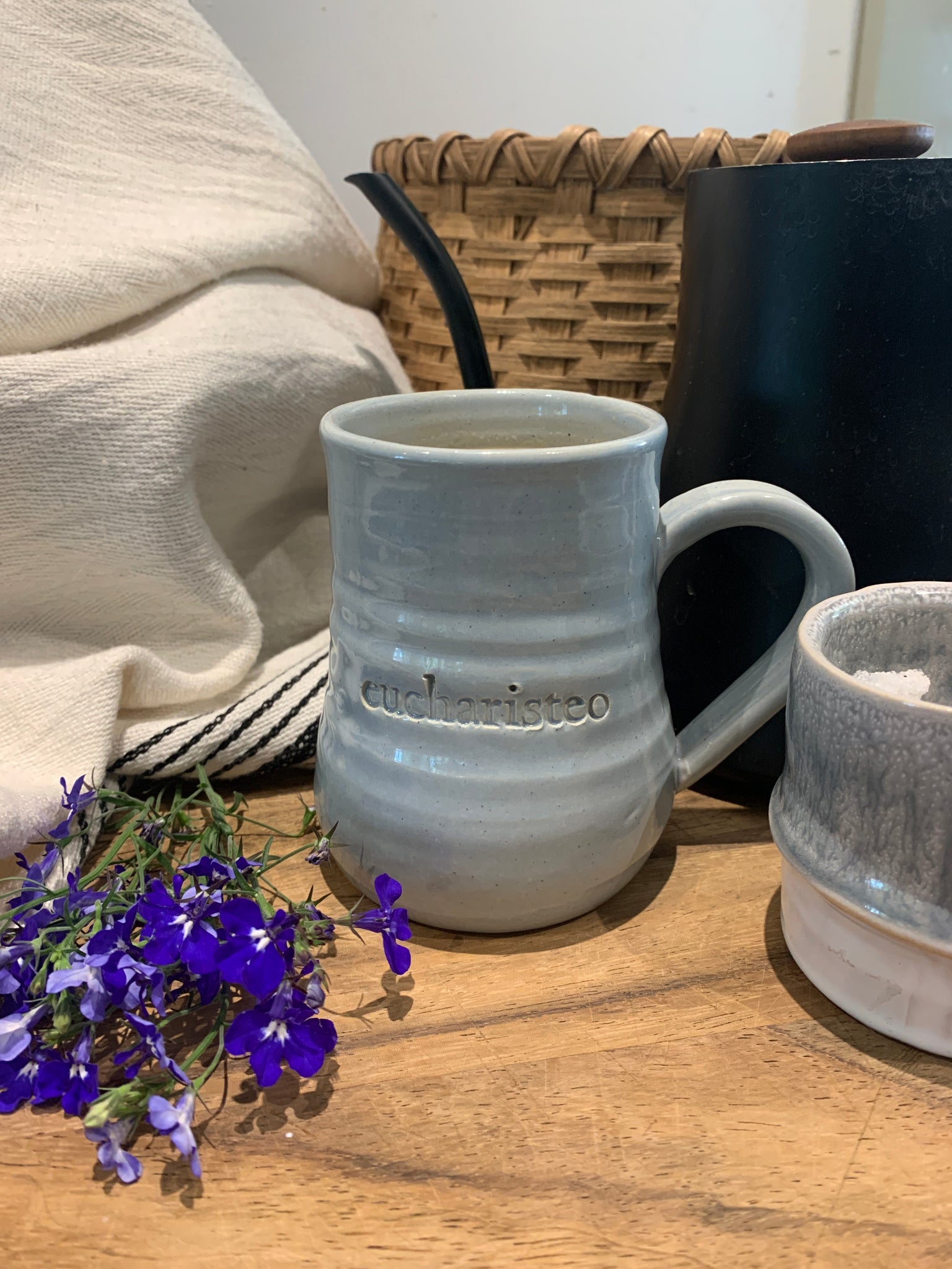 Three Gratitude Mugs with unique designs and glaze colors: "eucharisteo" with cloud blue glaze on white clay, "give thanks always" with darker blue glaze and ochre undertones, and "be still" with variegated gray glaze on red clay. Elegant shape, slight ripples, curved handles. Perfect for reflection and gifting.